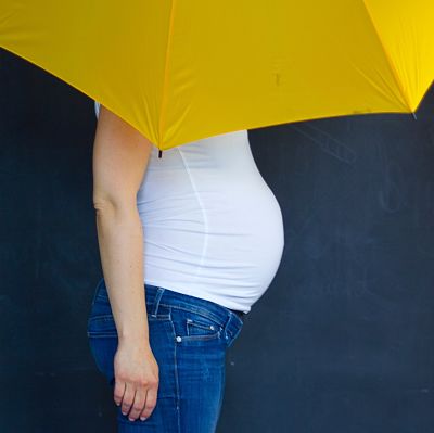 symbolfoto frau in weissem shirt und blauen jeans mit grossem babybauch, oberkörper teilweise abgedeckt durch einen gelben regenschirm
